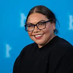 A dark-haired, smiling woman with long, dangling earrings looks right toward the camera