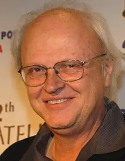 A profile image of Dennis Muren. A middle-aged, balding Caucasian male with short-white hair on the sides of his head. He is wearing dark glasses and looking towards the camera with a smile.