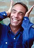 Derek Jarman wearing a denim shirt, smiling and putting hands against his ears