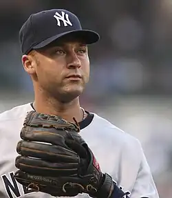 Derek Jeter wearing a navy hat and gray baseball uniform with a black glove stares into the distance.