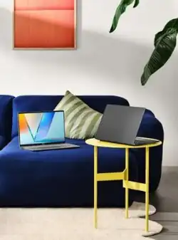 Open ASUS Vivobook on a blue couch displaying a colourful “V” wallpaper; another closed ASUS Vivobook sits on a small yellow table beside it, with a houseplant and abstract wall art in the background.