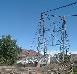 A view of a cable suspension bridge with one metal tower and a wooden deck. The second tower is partially obscured by a cottonwood tree.