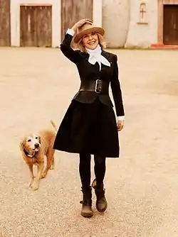 A smiling woman in a black belted dress, white scarf, and wide-brimmed hat walks with a golden dog across a sunlit courtyard