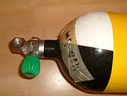 The shoulder of a scuba cylinder and a pillar valve are shown. The cylinder is yellow with black and white quartered shoulder, there is a tape stuck to the shoulder indicating maximum operating depth and the cylinder valve has a DIN connection opening directly above the neck thread, perpendicular to the cylinder axis, and a rubber knob on an orthogonal valve spindle in the right handed configuration.