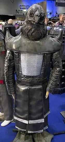 A grey, reptilian and amphibian humanoids. It has a turtle-like facial structure, with black and grey armor resembling that of a samurai.