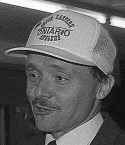 Boudria is a man in his late 50's, in a suit, with a moderately thin moustachee, shirt and tie, and wearing a light coloured ballcap that is too large for him, but not askance, reading "Eastern Ontario Anglers"