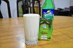A glass of white carbonated liquid on a wooden table. Next to the glass are a cardboard milk box and plastic bottle of Sprite soda.