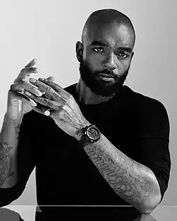 Portrait of Samuel Ross wearing a dark jumper, prominent wristwatch and pinkie ring; the tattoos on his forearms clearly visible.