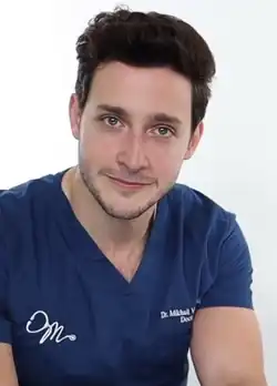 Headshot of Mike Varshavski. He is a strong, white man with dark brown hair, a stubble beard and moustache, and green eyes. He is wearing a T-shirt resembling medical scrubs with his name and logo, consisting of a stethoscope in the shape of the letter M.