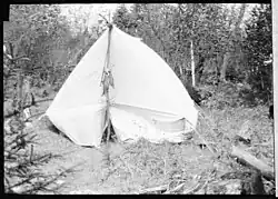 View of a camp site, with a white tent, showing a string of fish and a fly fishing rod hanging from the front tent pole.
