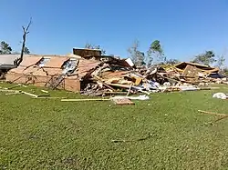 A red brick home surrounded by grass, seen destroyed. A debarked tree stands to its left.