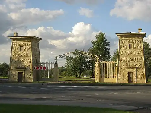 Stone Egyptian Revival cemetery gates with two towers covered in carved reliefs and an iron fence between them