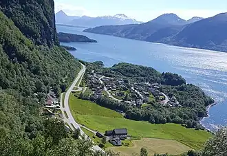 A small Norwegian village is depicted. We look down on it, surrounded by trees and other greenery. The village sits on the coast of a river, with islands in the background. Parallel to the village is a road. A cliff face sits to the left of the road.
