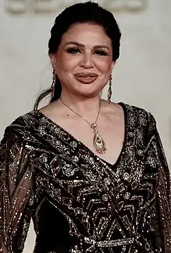 Elham Shahin wearing a black sequined outfit, grinning and appearing to be speaking while looking directly at camera