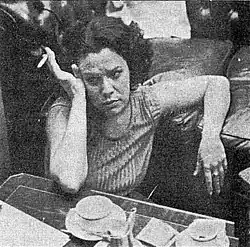 A dark-haired woman sitting a table, smoking a cigarette.
