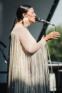 Elisapie seen from right profile, appearing to wear a traditional Indigenous outfit with matching earrings, standing onstage and speaking/singing into a microphone, both hands gesticulating