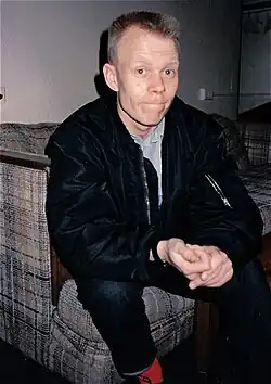 A young white man, sitting on the corner of a dilapited sofa, looking into the camera with an expression that is somewhere between a smile and a grimace. His brown hair is cut short and he's clasping his hands in front of him.