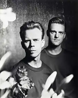 A black and white studio picture of Vince Clarke and Andy Bell. Both have short dark hair and are looking into the camera, with out of focus flowers in the foreground.