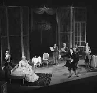 Black and white photograph of several people in eighteenth-century garb arranged on a stage. Near the centre, a man is singing with an arm outstretched toward a seated woman in an elegant dress.
