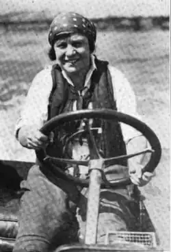 A smiling white woman wearing trousers and a headscarf, at the wheel of a speed boat