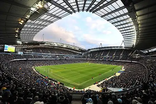 A football stadium as viewed from one corner. The photograph is a fish-eye paorama taken during a game against a blue sky. The stadium is full of fans on all four sides of the pitch, with large stands stretching up below an arched roof.