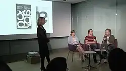A group of three seated people, one woman and two men, in a dark auditorium and a projection screen in the background, with another woman standing in front of the screen on the left