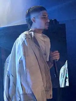 A photo of British rapper Fakemink performing on stage. He has a unique hairstyle with a mullet in the back and a fade on the sides.