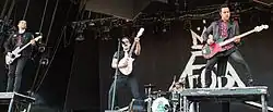 Four Caucasian men play musical instruments and sing into microphones on a stage.