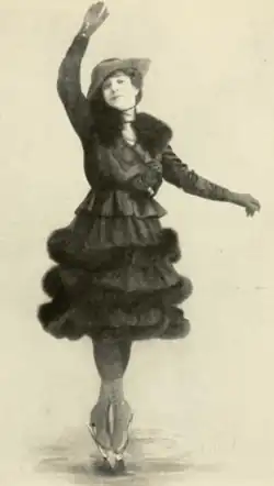 A white woman wearing a dark dress, in a figure skating pose