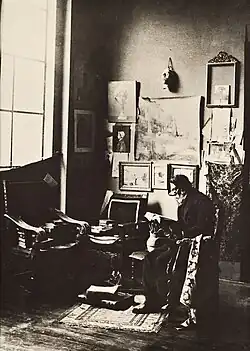 A seated man with a white beard reads a book at the corner of a studio.