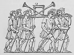 Roman soldiers carrying a platform (ferculum) adorned with trumpets and what looks like a table. They march in unison, wearing Roman attire, conveying a sense of order and ceremony.