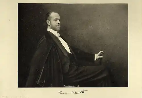 A bearded man in formal evening attire with a cape draped over his shoulders sits in profile, holding a cane