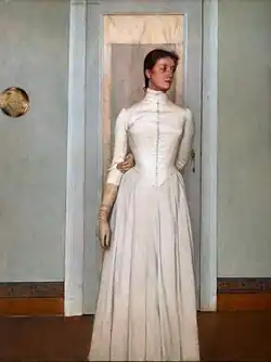 A woman with brown hair pulled back stands in a white room wearing a long white dress and tan gloves, her left arm bent behind her back with her hand resting in the crook of her right elbow as she gazes to the side