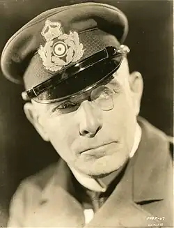 An actor wearing an Imperial German naval uniform and a monocle looks upwards and to his left, raising an eyebrow with a stern expression.