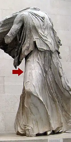A headless Greek statue of a woman in a long dress. Some colouration can be seen on its lower parts. An arrow has been digitally added about midway up its thigh