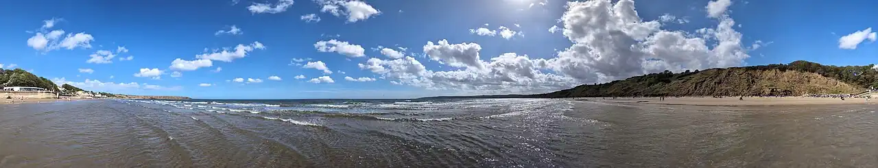 A sunny panoramic view of Muston Sands beach in Filey, showing a wide blue sky with white clouds, a calm sea, and people walking on the sand next to low, earthy cliffs.