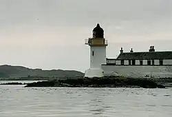 A small white lighthouse with a black canopy sits on a dark rock. There is a high white wall and a row of black and white houses to the right and dark hills at left in the distance.