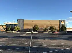 This photograph shows the east side of Flatirons Community Church in Lafayette, Colorado, including the elevated glass entry volume at the northeast corner, long banded brick wall panels with vertical metal fins, regularly spaced parking-lot lights and newly planted street trees, and a largely empty asphalt lot under clear morning light.