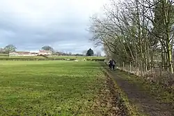 A field with a line of trees on the right hand side and a collection of farm buildings in the distance atop a gentle slope
