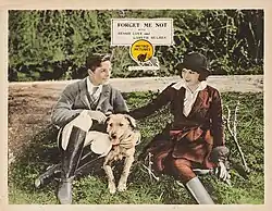 A hand-colored lobby card with a man and woman in riding clothes sitting on the grass with a dog between them
