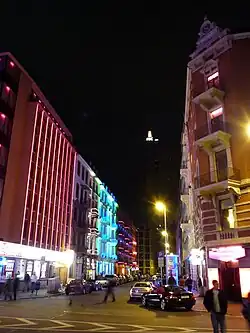 A city street at night, with many colourfully lit buildings. On the left is a large, red tower.