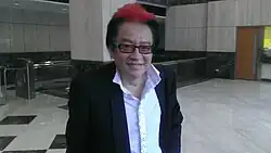 Frankie Kao wearing a white ruffled shirt and dark suit jacket, with a red blaze in his hair, grinning at camera