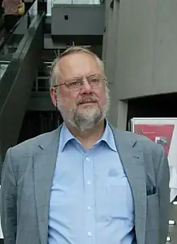 A bearded older man in a blue dress shirt and grey sports coat wearing glasses