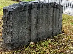 The image shows the Liberty Point Resolves Monument in Cross Creek, Fayetteville, North Carolina. The monument is a granite stone with a rounded top and jagged edges, standing on a grassy area. Centered on the smooth front face of the monument are the names of thirty-nine signers of the Liberty Point Resolves. At the top, the text reads “Declaration of Independence June 20, 1775,” The stone is dark gray and irregularly shaped on the sides and top. In the background, a metal fence lines a sidewalk and a road beyond it.