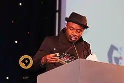 Man accepting award, wearing black hat and top.