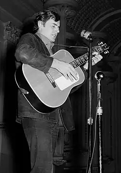 A man plays an acoustic guitar and sings into a microphone on a stand. He is shown in right profile, wearing dark clothes and looking downwards.