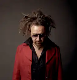 Gaudi wearing a black shirt and red jacket, facing camera with head tilted downward