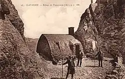 Three men stand outside a stone building in a pass between two rock features.