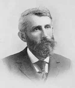 A man in his forties, facing right, with thick black hair, a beard, and a mustache. He is wearing a white shirt, black tie, and black jacket