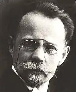 Black and white studio portrait of an unsmiling man wearing pince-nez, with short, dark hair brushed back, and a trimmed, greying beard.